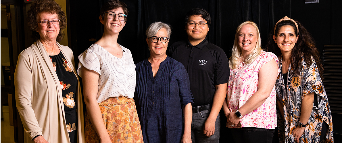 Six people stand side by side, smiling at the camera in an indoor setting with a dark backdrop. The group includes individuals of different ages and appearances, dressed in business-casual clothing. From left to right: a person in a light cardigan over a floral dress, a person in a white polka-dot blouse and floral skirt, a person with short gray hair wearing glasses and a navy blouse, a person in a black polo shirt with an 