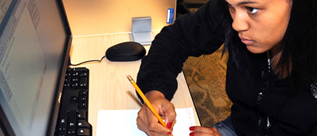 Person writing on a notepad while looking at a computer monitor at a desk, with a keyboard, pencil, and mouse visible.