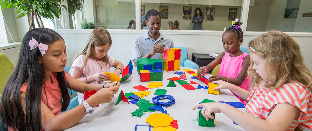 Five children and an instructor sit around a table, building colorful geometric structures with magnetic tiles in a bright classroom while collaborating on hands-on learning activities.