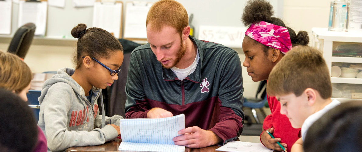 Teacher providing one-on-one support to a small group of elementary students seated around a classroom table, reviewing notes together during a lesson.